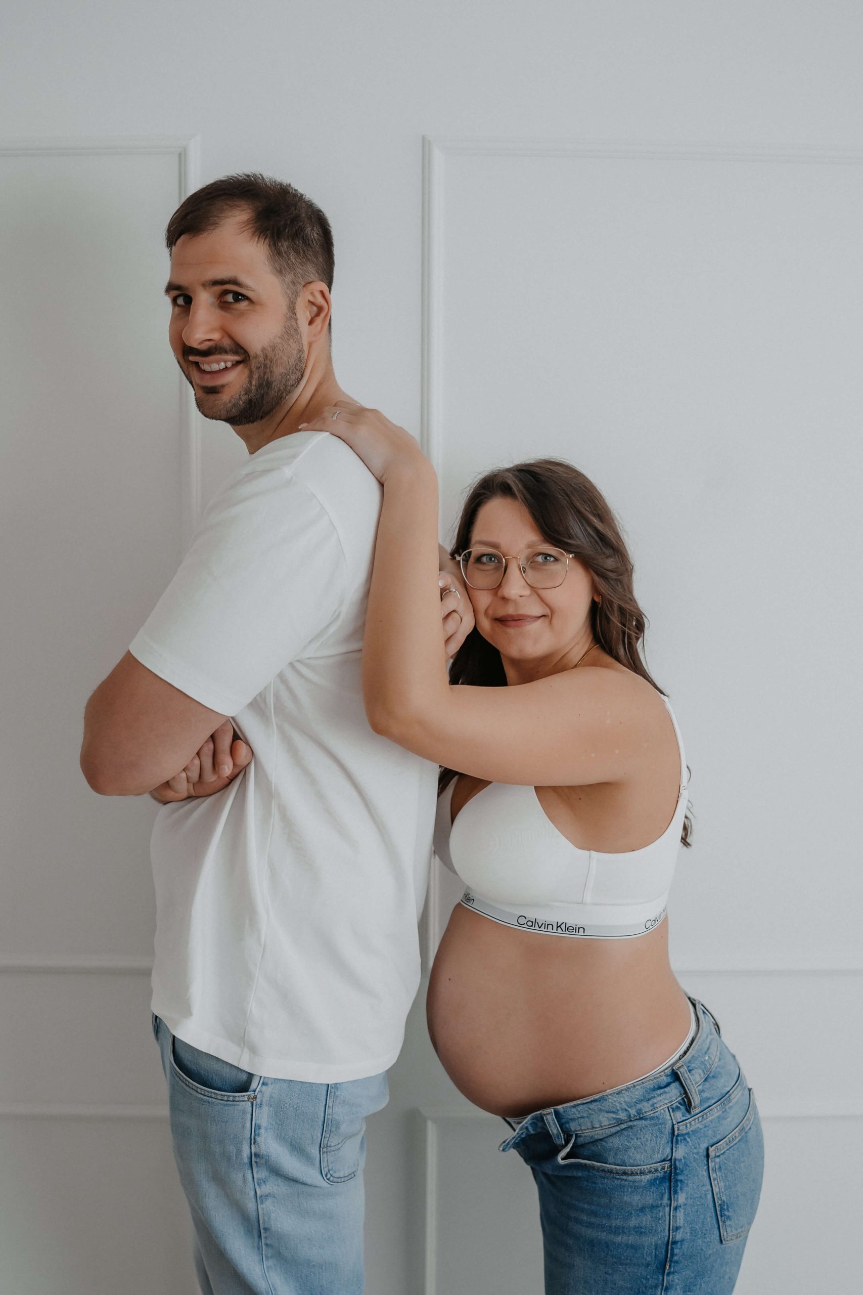 Schwangere Frau umarmt lächelnden Mann von hinten, beide tragen weiße Oberteile und Jeans, stehen vor hellem Hintergrund. © Augenblick Photography