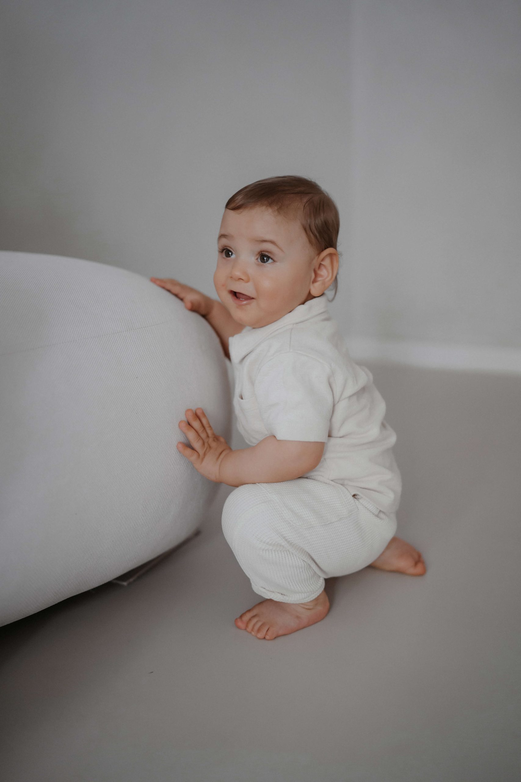 Ein Baby in cremefarbener Kleidung kniet und stützt sich lächelnd an einem großen, weißen Polster in einem neutralen Raum ab. © Augenblick Photography