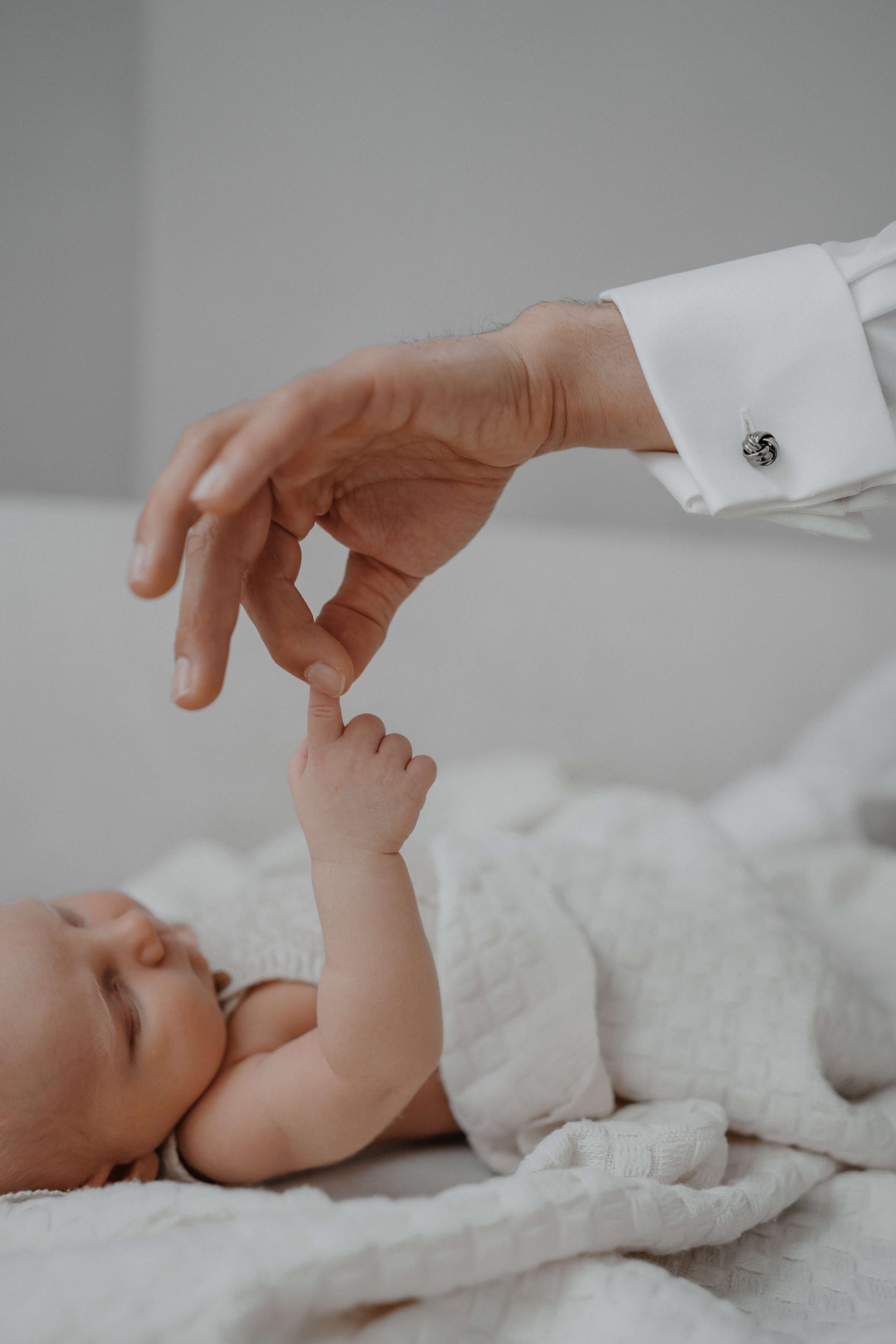 Ein Baby hält den Finger eines Erwachsenen, der einen weißen Hemdärmel trägt, während es auf einer weißen Decke liegt. © Augenblick Photography