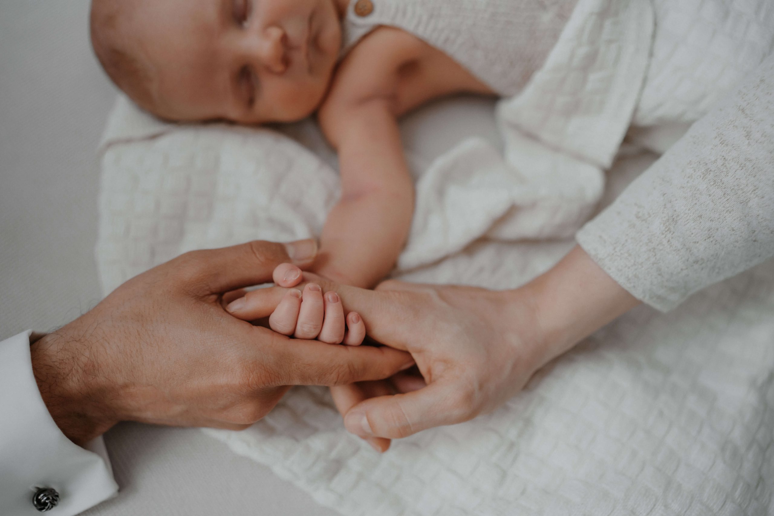 Ein schlafendes Baby hält die Hände von zwei Erwachsenen fest, warmes, liebevolles Zusammensein, sanftes natürliches Licht, weiche Decke im Hintergrund. © Augenblick Photography